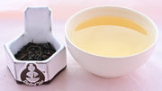 A side-by-side comparison of Wooricha leave and steeped tea. On the left, the leaves are narrow and curving, and a dark black color. On the right, the steeped tea is a rich yellow color.