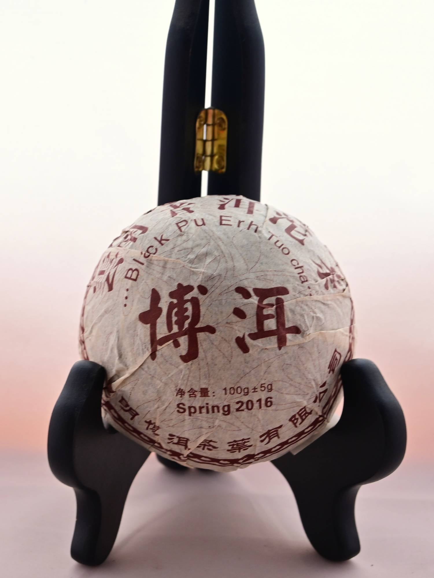 The Xiang Wei Tuo Cha dome rests in a black book holder. It is wrapped in white tissue paper with red designs of flowers in the background. Chinese letters circle the outside, with English translations inside reading: "Black Pu Erh Tuo Cha. Spring 2016. 100 grams."