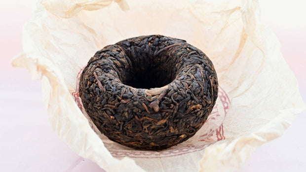 The Xiang Wei Tuo Cha dome rests upside down against its creme tissue paper wrapper. The leaves curl toward the hollowed inside of the dome, and range in color from black to dark orange.