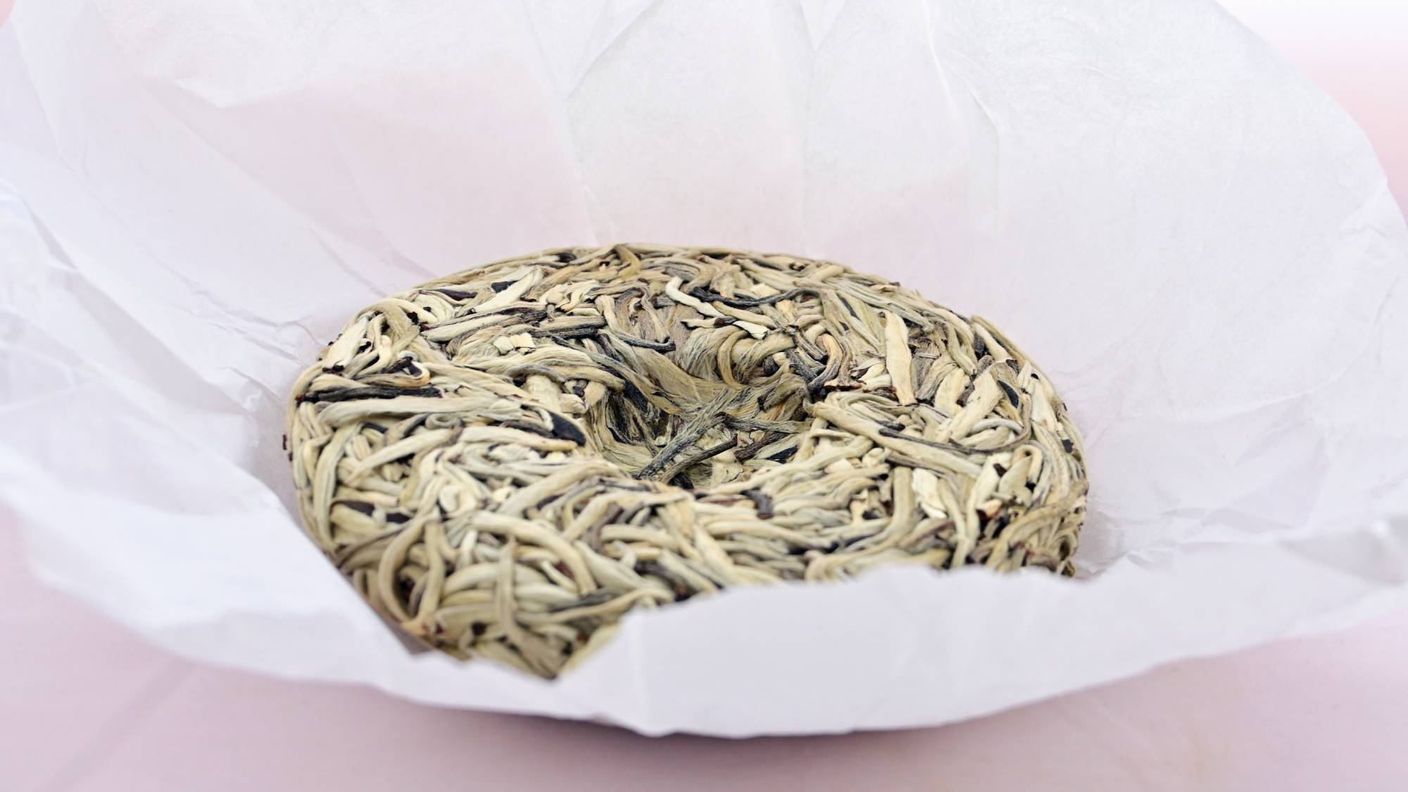 The Yin Zhen Bing Cha disk rests in the middle of its open tissue paper wrapper. The leaves are all white with a creamy golden sheen, and have been pressed together into a circle with a spherical cut out in the center.