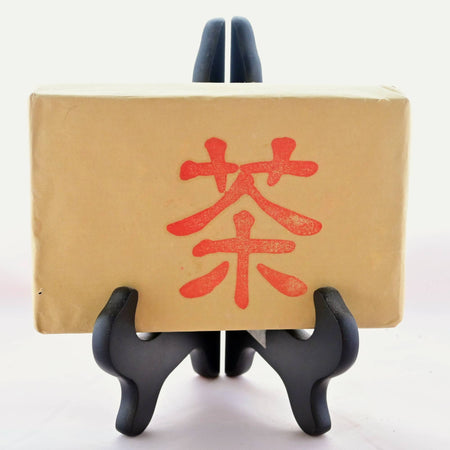 The Zhuan Cha tea brick is wrapped in brown paper and resting in a black book stand. On the front is a single Chinese character stamped in red ink.