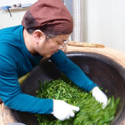 An image captures the pan-roasting step of Nok Cha processing. The processor is in a blue shirt, and leans over a wok-style pan while swirling their hand through vibrant green leaves.