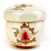 A close-up of the traditional Nok Cha drinking vessel, a single-serving cup with removable filter. This one has a cream base color, with leaf and flower designs in brown and red.