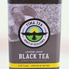 A triangular silver tea tin holds Black Rwandan tea. At the top is an image of a tea-picker smiling into the camera with a basket on their back. In the center, a logo featuring a bird with wings spread flying upward toward the sun is surrounded by words "Tima Tea. Spirit of Resilience." Below, the tin features other descriptive messaging: "Loose leaf black tea direct from Rwanda. Get loose... step out of the bag! Net weight is 2 ounces or 23 servings."